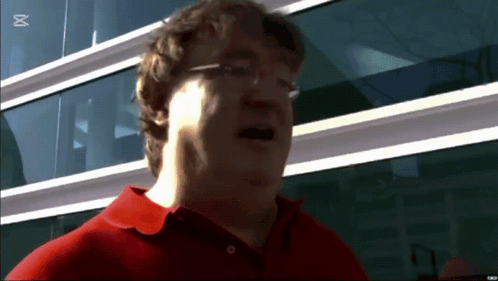 A Man Wearing Glasses And A Red Shirt Stands In Front Of A Building With The Letter M On The Bottom Right GIF
