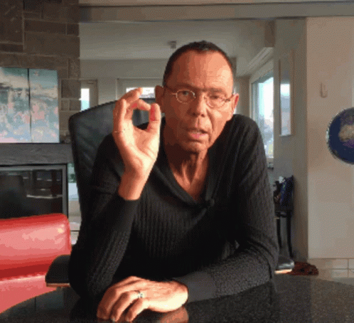 A Man Wearing Glasses And A Ring Sits At A Table With His Hands Folded GIF