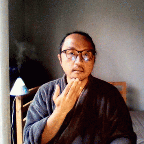 A Man Wearing Glasses And A Robe Looks At The Camera With His Hand On His Chin GIF