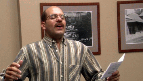 A Man Wearing Glasses And A Striped Shirt Sings Into A Microphone GIF