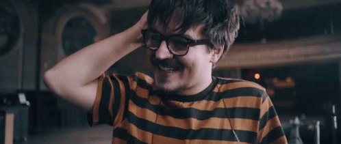 A Man Wearing Glasses And A Striped Shirt Smiles With His Hand In His Hair GIF