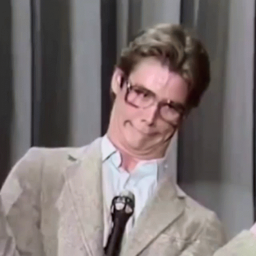 A Man Wearing Glasses And A Suit Is Standing In Front Of A Microphone Making A Funny Face . GIF