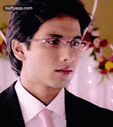 A Man Wearing Glasses And A Suit Is Standing In Front Of Flowers . GIF