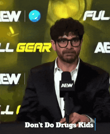 A Man Wearing Glasses And A Suit Stands In Front Of A Sign That Says Aew GIF