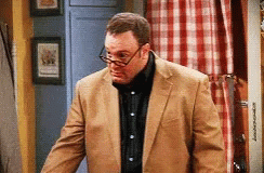 A Man Wearing Glasses And A Tan Suit Is Standing In A Room GIF