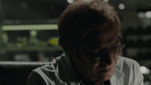 A Man Wearing Glasses And A White Coat Looks Down GIF