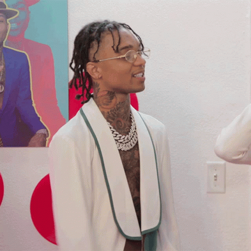 A Man Wearing Glasses And A White Jacket Stands In Front Of A Painting GIF
