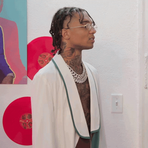 A Man Wearing Glasses And A White Robe Is Standing In Front Of A Wall With Red Records On It GIF
