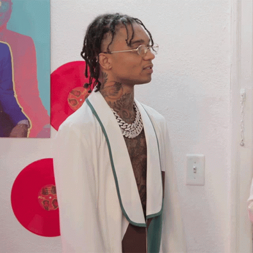 A Man Wearing Glasses And A White Robe Stands In Front Of A Wall With Red Records GIF