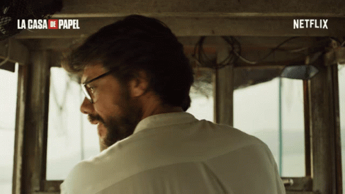 A Man Wearing Glasses And A White Shirt Is Sitting In Front Of A Sign That Says La Casa De Papel And Netflix GIF