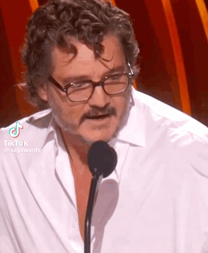 A Man Wearing Glasses And A White Shirt Is Speaking Into A Microphone . GIF