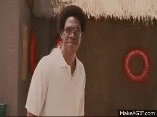 A Man Wearing Glasses And A White Shirt Is Standing In Front Of A Thatched Roof And Smiling . GIF