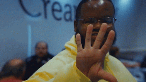 A Man Wearing Glasses And A Yellow Jacket Holds Up His Hand In Front Of A Wall That Says Crea GIF