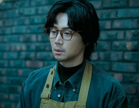A Man Wearing Glasses And An Apron Stands In Front Of A Black Brick Wall GIF