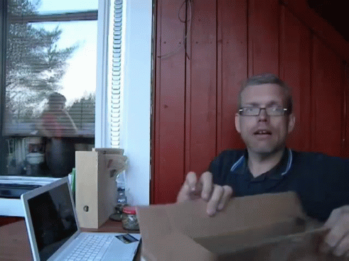 A Man Wearing Glasses Holds A Cardboard Box In Front Of A Laptop Computer GIF
