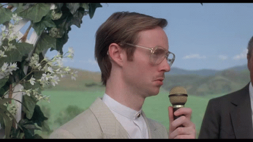 A Man Wearing Glasses Holds A Microphone In His Right Hand GIF