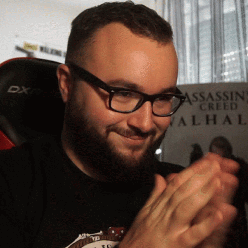 A Man Wearing Glasses Is Clapping In Front Of A Assassin Creed Valhal Poster GIF