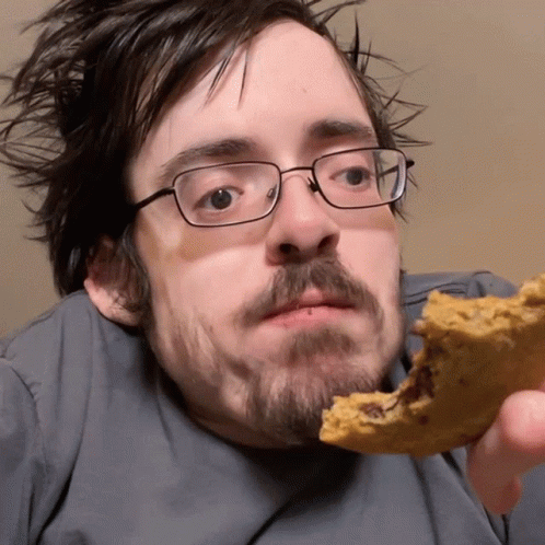A Man Wearing Glasses Is Eating A Cookie With A Bite Taken Out Of It GIF