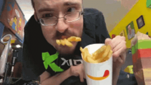 A Man Wearing Glasses Is Eating French Fries From A Cup With A Smile On It GIF