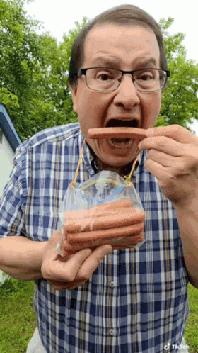 A Man Wearing Glasses Is Eating Hot Dogs From A Bag GIF