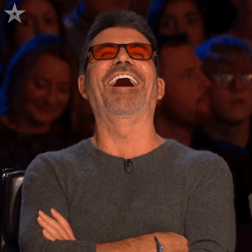 A Man Wearing Glasses Is Laughing While Sitting In Front Of A Crowd GIF