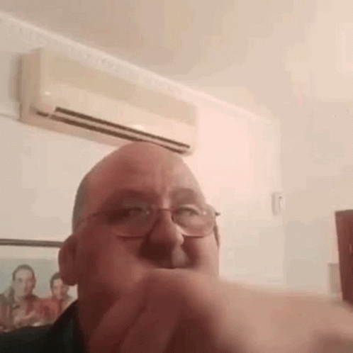 A Man Wearing Glasses Is Making A Funny Face In Front Of An Air Conditioner In A Room . GIF