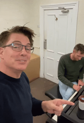 A Man Wearing Glasses Is Pointing To Another Man Playing A Piano GIF