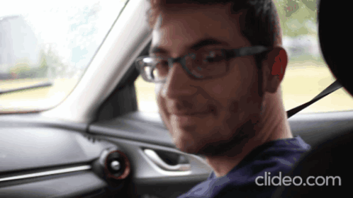 a man wearing glasses is sitting in the driver 's seat of a car with clideo.com displayed on the bottom