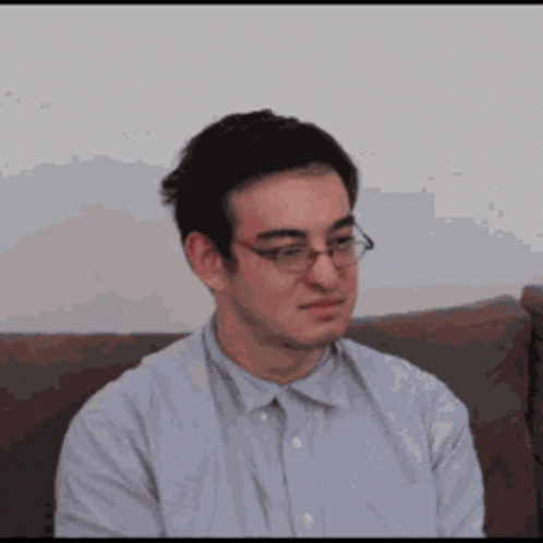 A Man Wearing Glasses Is Sitting On A Couch And Looking At The Camera . GIF