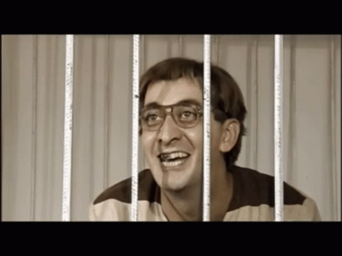 A Man Wearing Glasses Is Smiling Behind Prison Bars GIF