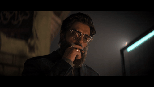 A Man Wearing Glasses Is Smoking A Cigarette In Front Of A Wall With Arabic Writing On It GIF