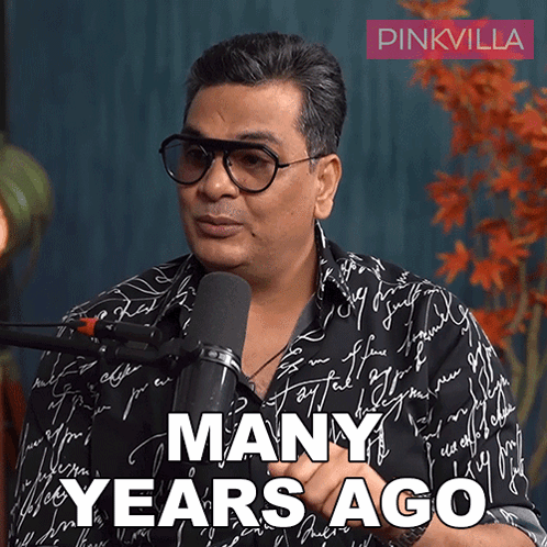 A Man Wearing Glasses Is Talking Into A Microphone With The Words Many Years Ago Below Him GIF