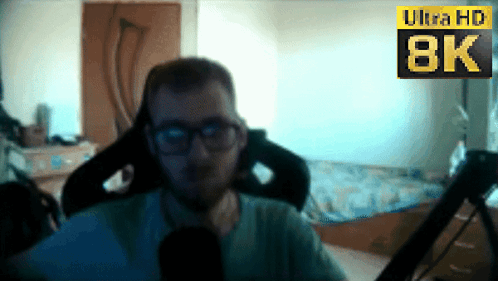 A Man Wearing Glasses Sits In Front Of An 8K Sign GIF