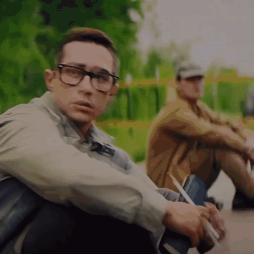 A Man Wearing Glasses Sits Next To Another Man Who Is Smoking A Cigarette GIF