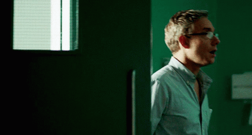 A Man Wearing Glasses Stands In Front Of A Green Wall GIF