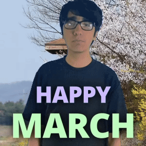A Man Wearing Glasses Stands In Front Of A Sign That Reads Happy March GIF