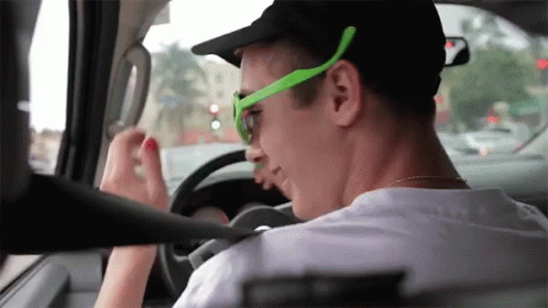 A Man Wearing Green Sunglasses And A Black Hat Driving A Car GIF