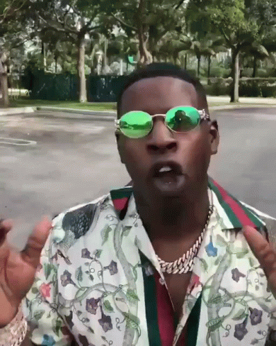 a man wearing green sunglasses and a floral shirt stands in a parking lot