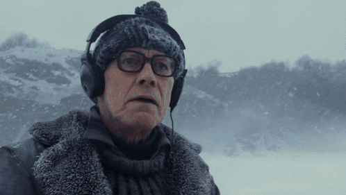 A Man Wearing Headphones And A Beanie Looks At The Camera In The Snow GIF