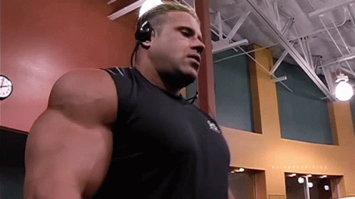 A Man Wearing Headphones And A Black Tank Top Stands In Front Of A Clock GIF