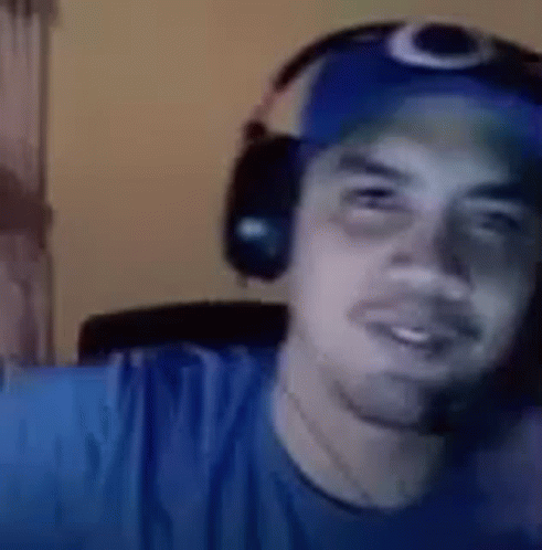 A Man Wearing Headphones And A Blue Hat Is Looking At The Camera . GIF