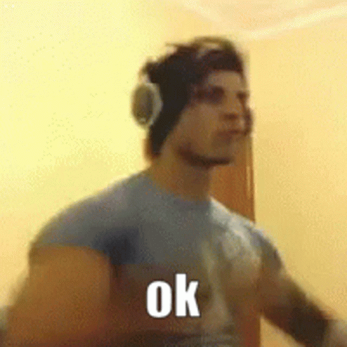 A Man Wearing Headphones And A Blue Shirt Is Standing In Front Of A Wall And Says Ok . GIF