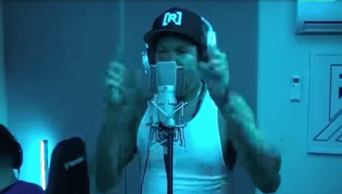A Man Wearing Headphones And A Hat With The Letter R On It Is Singing Into A Microphone GIF