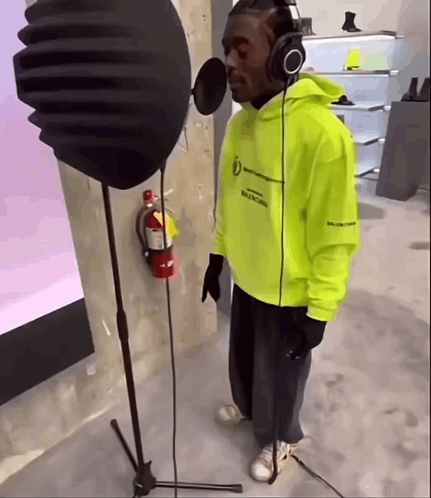 A Man Wearing Headphones And A Neon Yellow Hoodie Sings Into A Microphone GIF