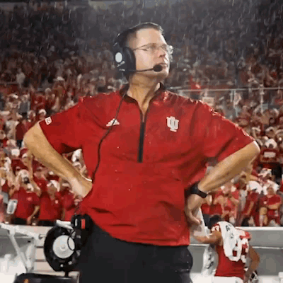 A Man Wearing Headphones And A Red Shirt With The Letter I On It Stands On A Football Field . GIF