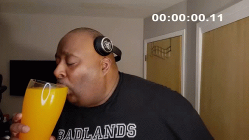 A Man Wearing Headphones And A Shirt That Says Badlands Drinks A Glass Of Orange Juice GIF