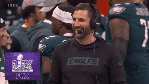 A Man Wearing Headphones And A Shirt That Says Eagles Laughs In Front Of Football Players GIF