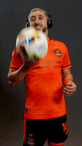A Man Wearing Headphones Holds A Soccer Ball While Wearing An Orange Anderson Career Center Jersey GIF