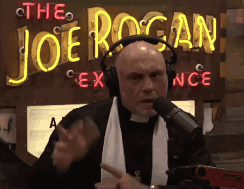 A Man Wearing Headphones Is Talking Into A Microphone In Front Of A Neon Sign That Says The Joe Rogan GIF