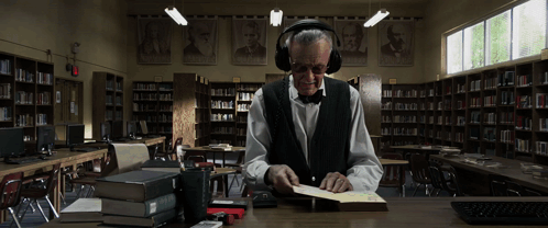 A Man Wearing Headphones Sits At A Desk In A Library Reading A Book GIF